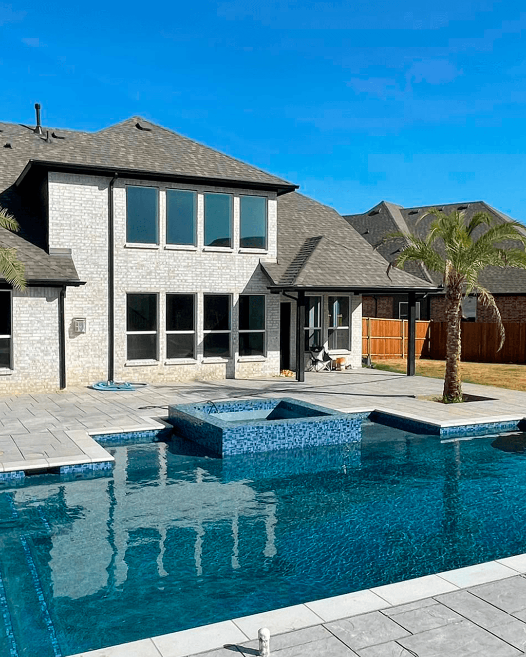 Luxury backyard pool in Frisco, Texas, at dusk featuring a rock grotto waterfall, integrated spillover spa, fire bowls, blue LED lighting, and a tanning ledge with lounge chairs.
